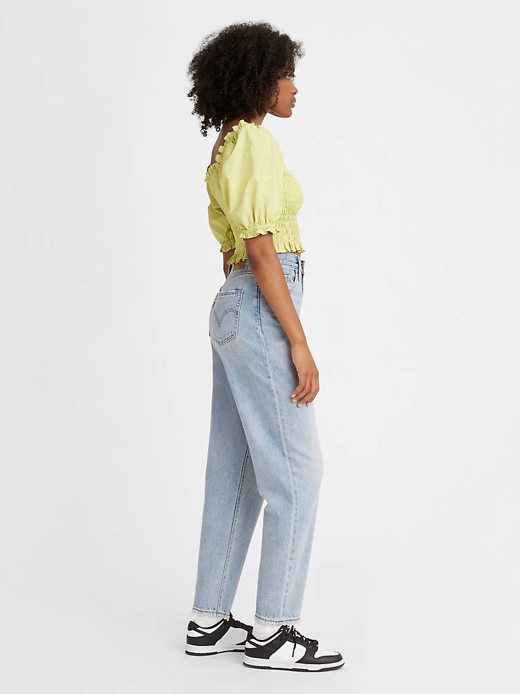 Women is wearing Levi's high loose tapper fit jeans. These Jeans are light blue color, high waistes and ripped. Jeans are paired with light green cropped top and black sneakers. The photo is taken from the side. (7721828384976)