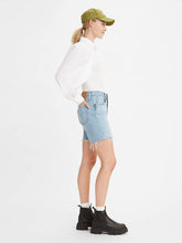 Load image into Gallery viewer, Women is wearing Levi&#39;s 501 original mid thigh shorts in light wash color. Jean shorts are styled witha white shirt and black boots. Jean shorts feature high waist, bermuda lenght and classic straight fit. The photo demostrates jean shorts from the side. (7721852010704)
