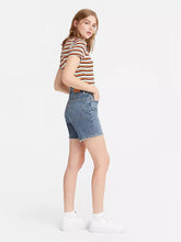 Load image into Gallery viewer, Woman is wearing Levi&#39;s mid thigh shorts. Jean shorts feature high waist, mid thigh length and cut off hem. The photo demonstartes the model from the side. (7721825140944)
